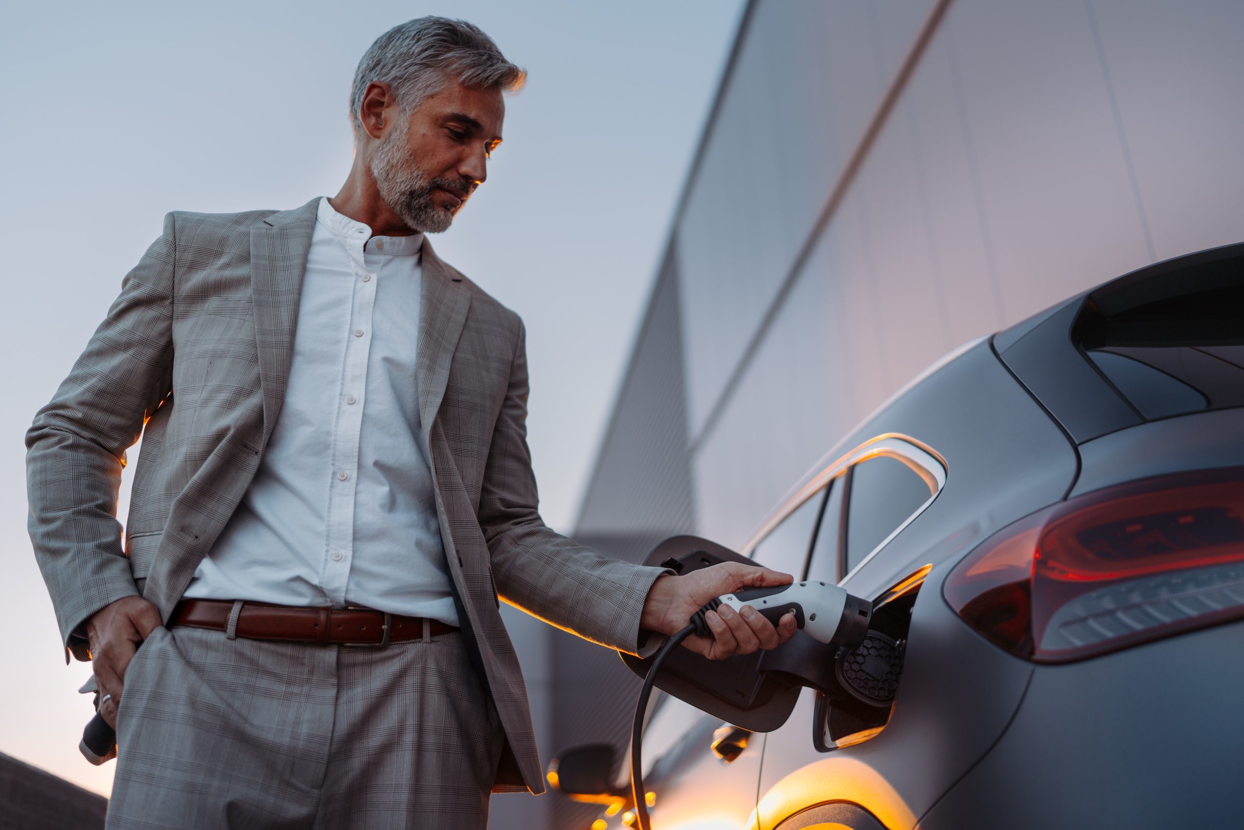 Businessman, Holding Power Supply And Charging His Electric Car During Sunset. Concept Of Ecology Tranport.