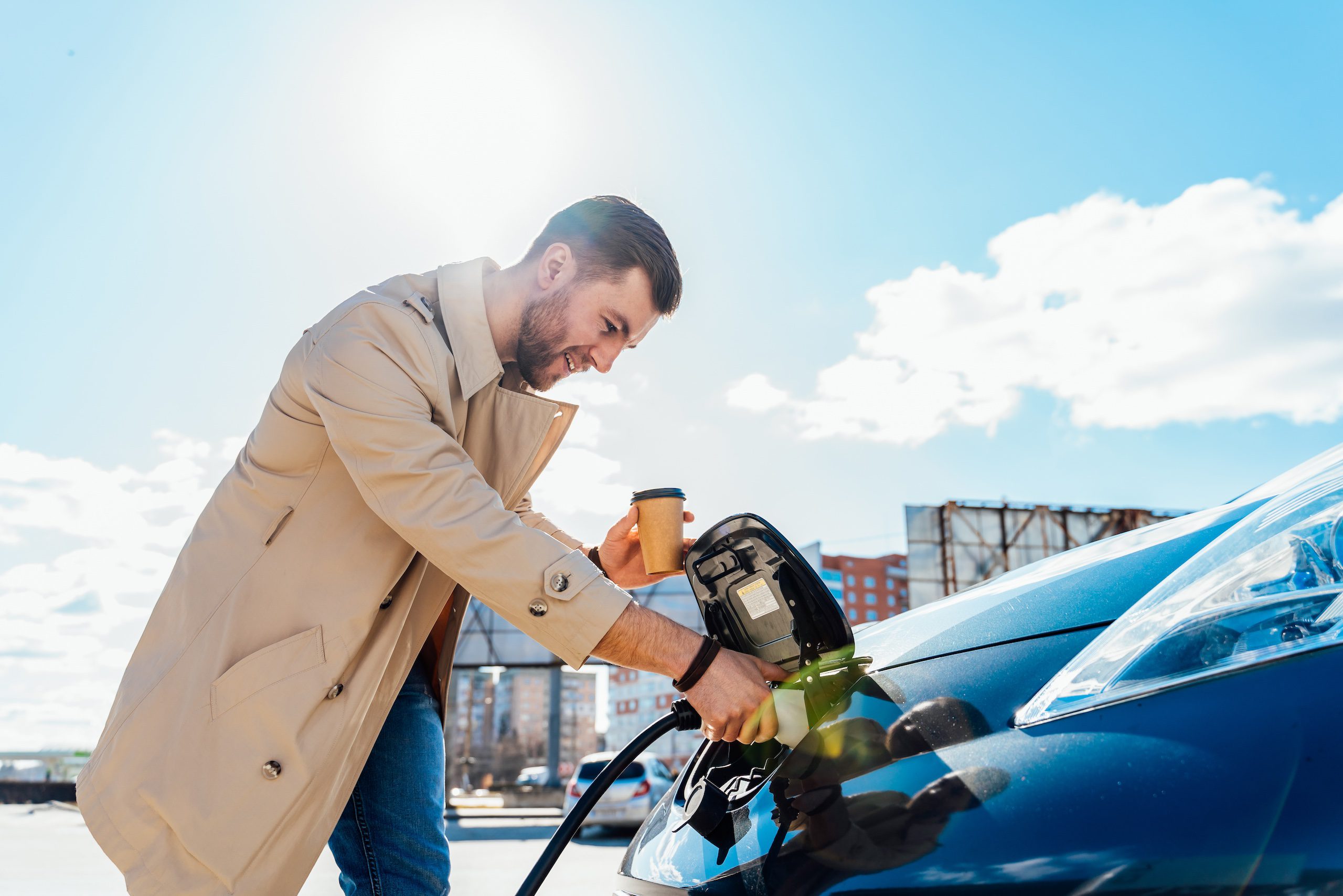 Stylish Man With Coffe Cup In Hand Inserts Plug Into The Electric Car Charging Socket